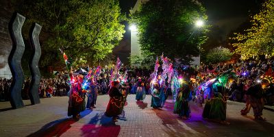 Multiple dancers, wearing colourful, light-up costumes, dancing in front of a wavy statue outside at night, surrounded by a circle of viewers sat on stone walls.