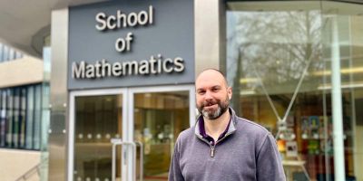 Person standing outside the entrance to the School of Mathematics building at the University of Leeds, with the building’s glass doors and large sign visible in the background.