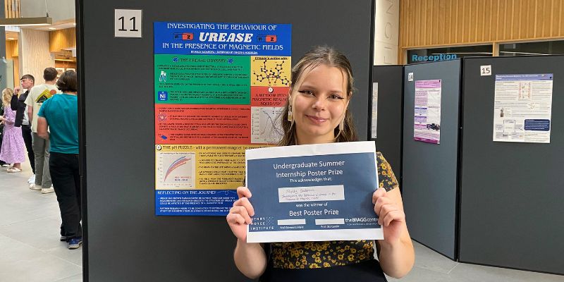 A cropped photo of Nelli Sadarova holding her 1st prize certificate, while stood in front of her winning poster design.