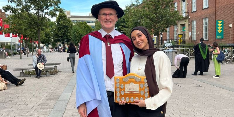 Farida stood with a professor, holding her Simon Vickers award.