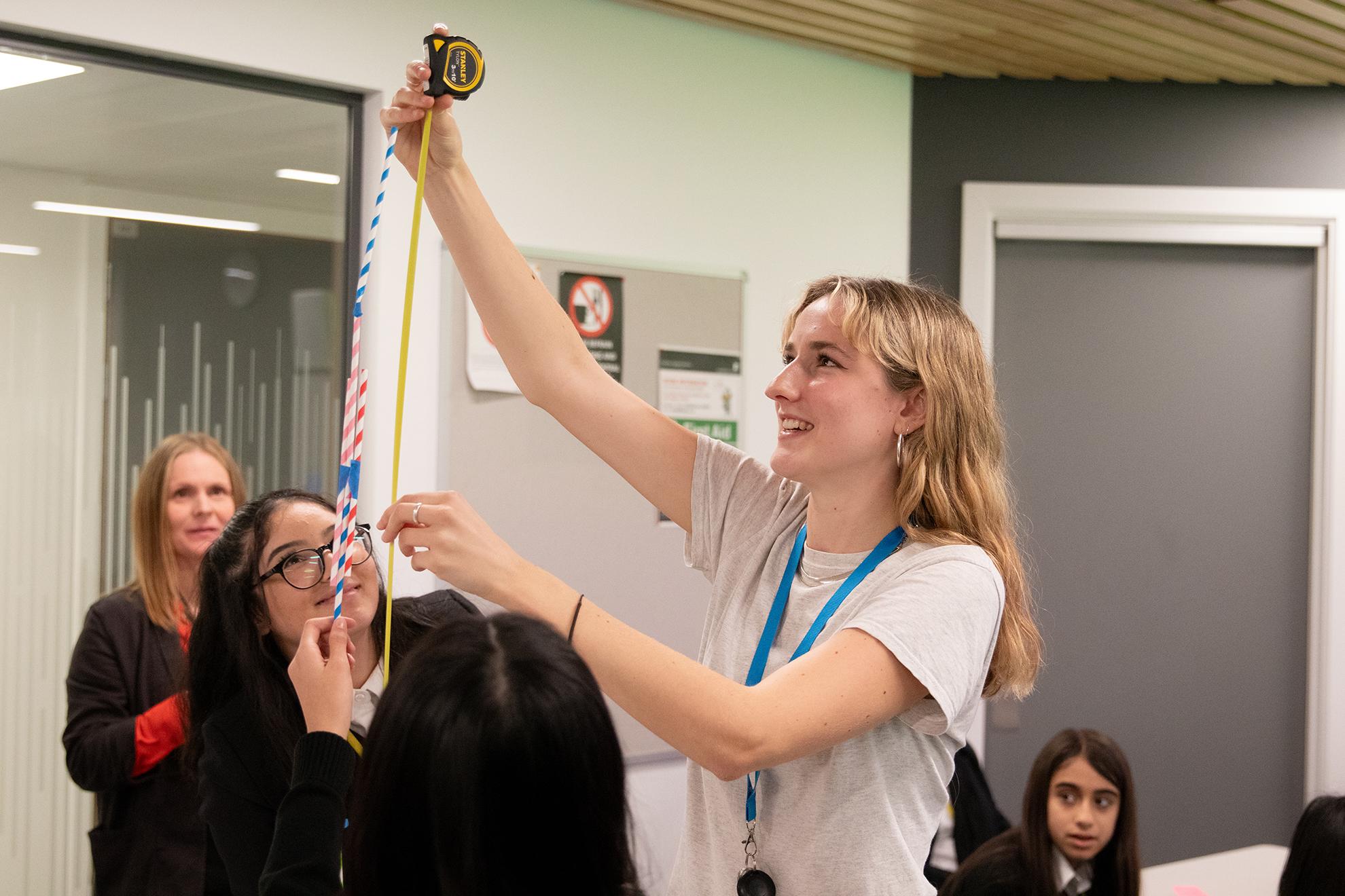 Someone measuring a tower made from straws, using a tape measure, while surrounded by young students.