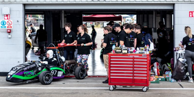 A photograph of the Gryphon Racing team, in the garage at Silverstone, pushing the car out on to the track. The team are in their uniform and one student is getting some tools from a red tool box on wheels.