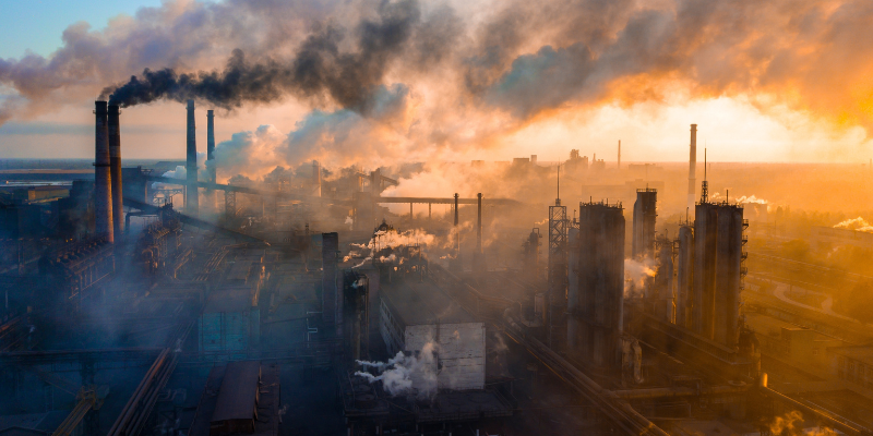 An industrial area with multiple factories emits smoke and steam as the sun sets, casting an orange hue over the scene.