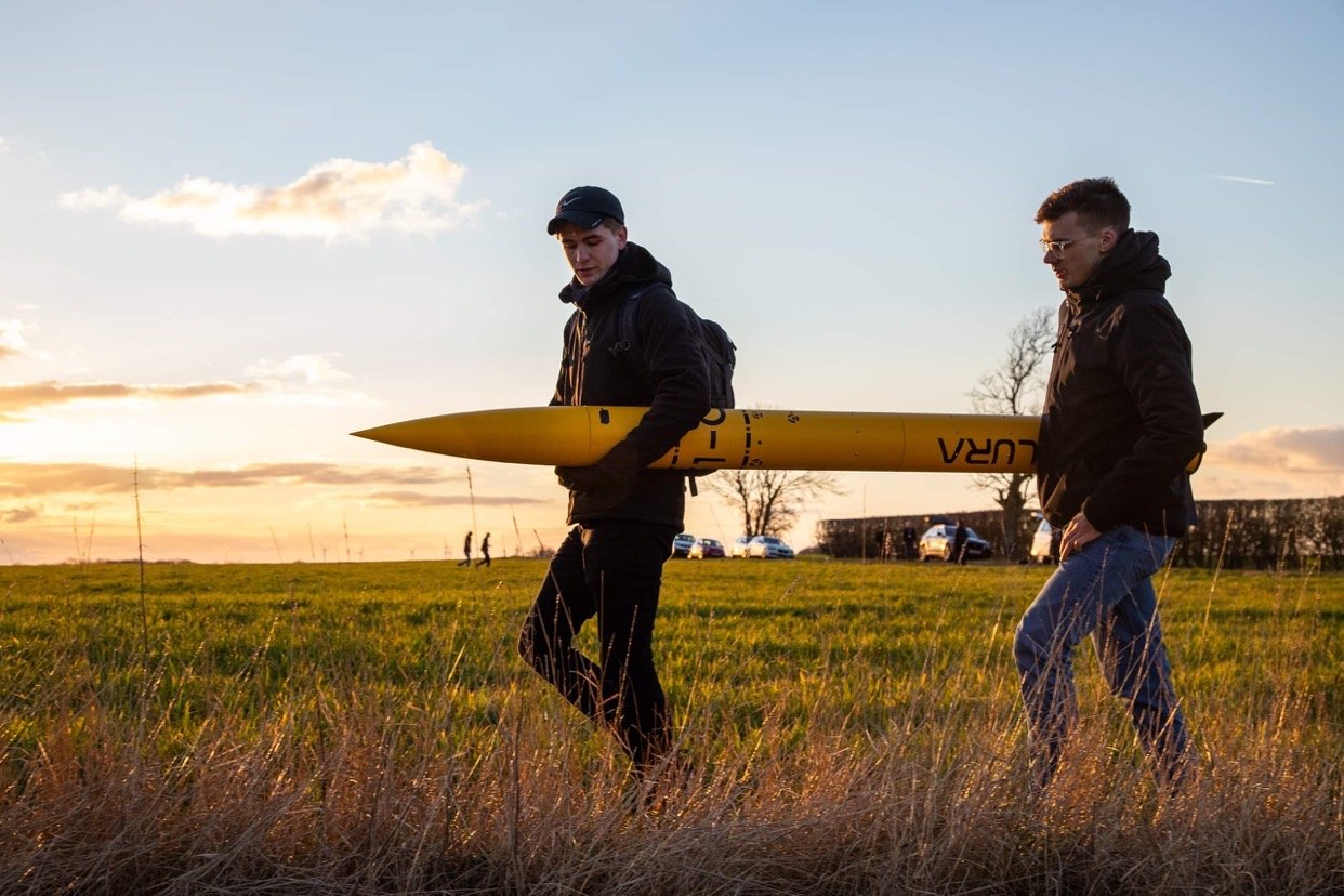 A photograph of two students walking across a field carrying a rocket between them.
