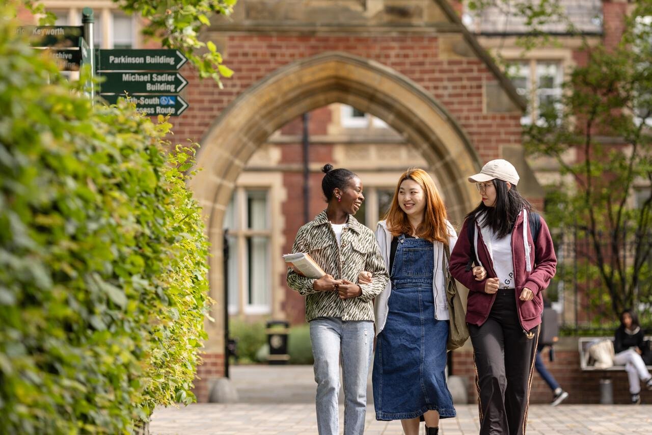 Three students walk together, smiling and chatting, on a sunny campus. They are near a brick archway with directional signs, surrounded by greenery.