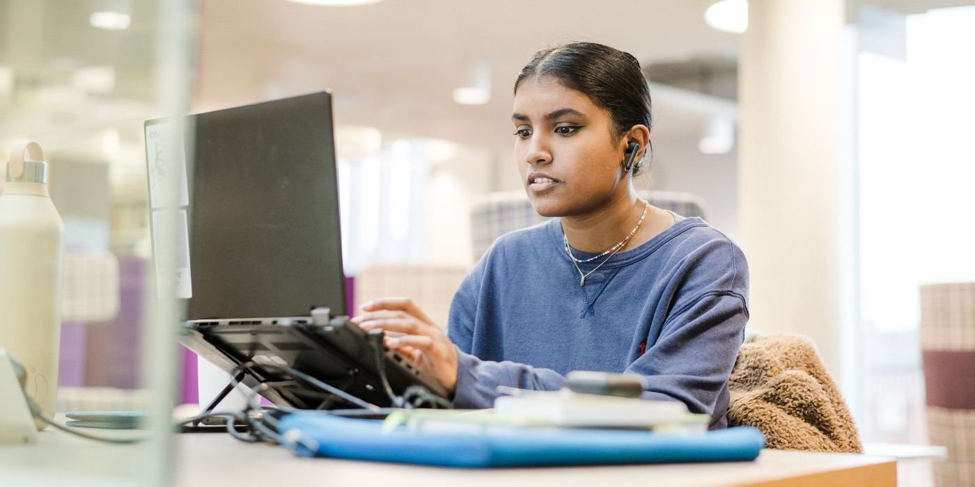 A student working on a laptop in a library, the student is in focus and the background is blurred.