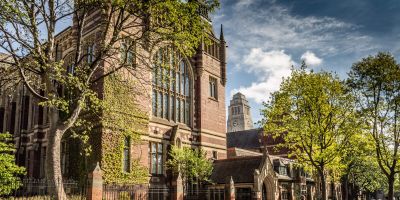 Historic red-brick building at the University of Leeds surrounded by trees under a partly cloudy sky.