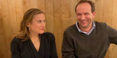 Two people sitting indoors against a wooden panel wall, dressed in smart casual clothing.