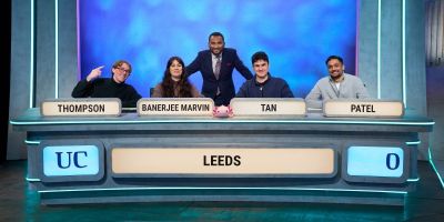 The 2024 University Challenge Leeds team, on set and sat behind their desk, with host Amol Rajan standing behind them in the centre of the shot.