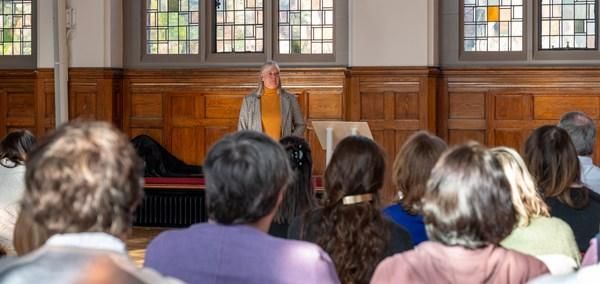 A person stands at a podium addressing an audience seated in a room with wood-panelled walls and stained glass windows.