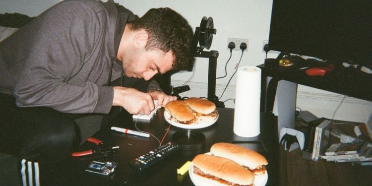 A photograph of Ryan Jamal working on a circuit board at home on a coffee table, with a sandwich next to him.