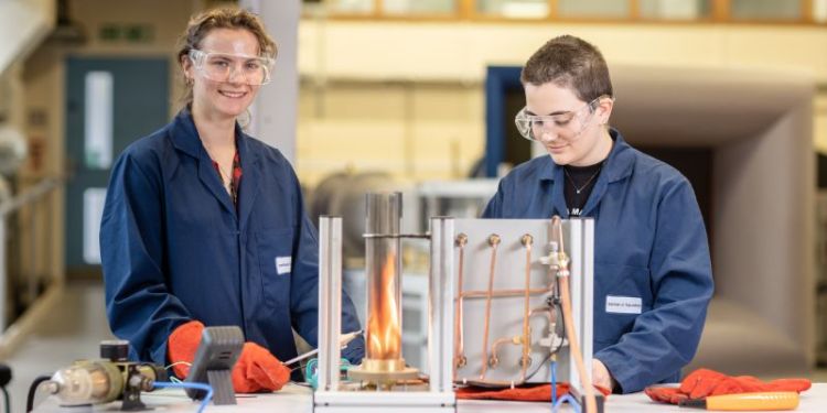 Two students working on a combustion workbench in the School of Mechanical Engineering, at the University of Leeds. The students are in blue protective coats and gloves and are performing an experiment with a flame.