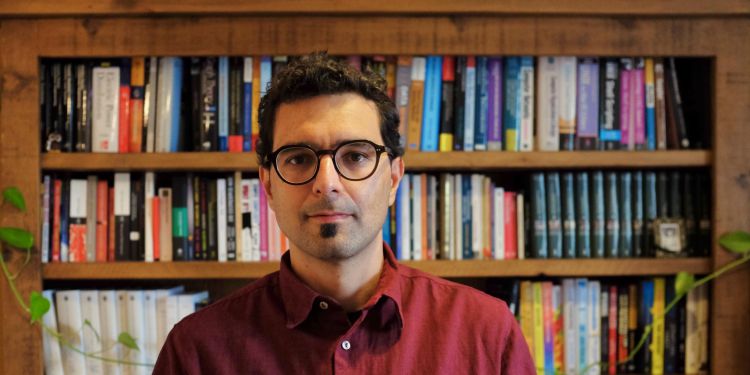 Picture of a man wearing glasses and a red shirt standing in front of a book sheld