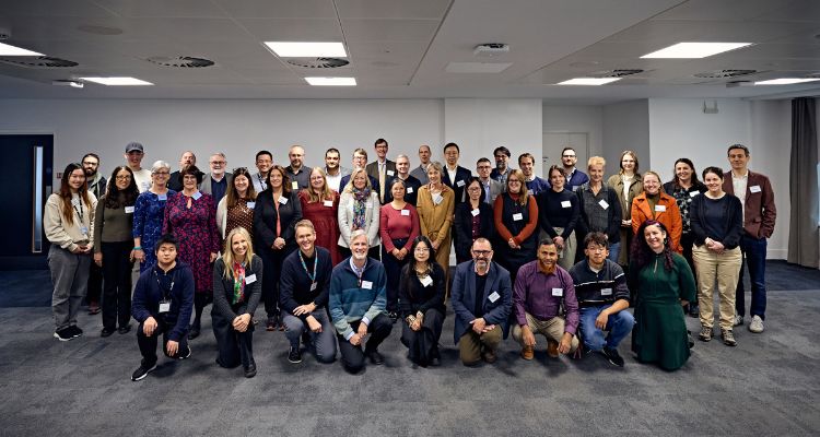 “Group photo of conference attendees standing and kneeling in rows inside a modern meeting room with grey carpet and white walls. Participants are wearing name badges and a mix of business and casual attire, with some smiling towards the camera.”
