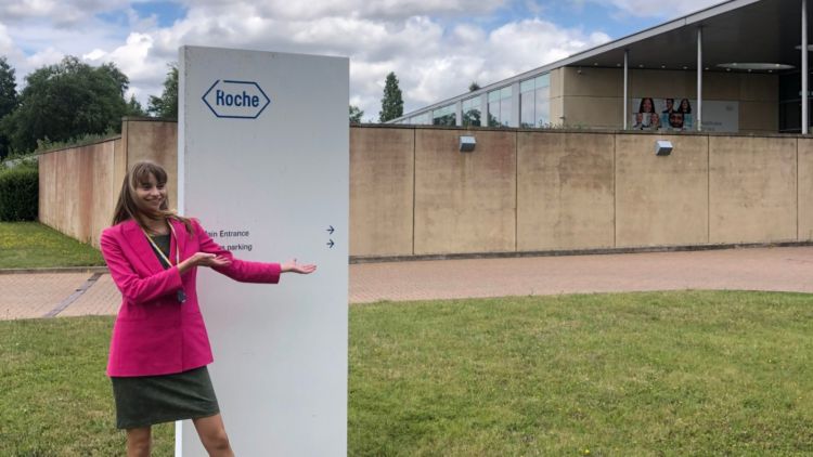 Natalja nassonova in a pink suit at her year in industry placement. She is standing by a white sign, pointing at her office.