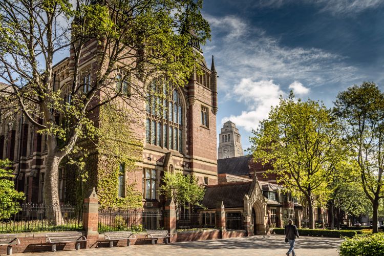 Historic red-brick building at the University of Leeds surrounded by trees under a partly cloudy sky.