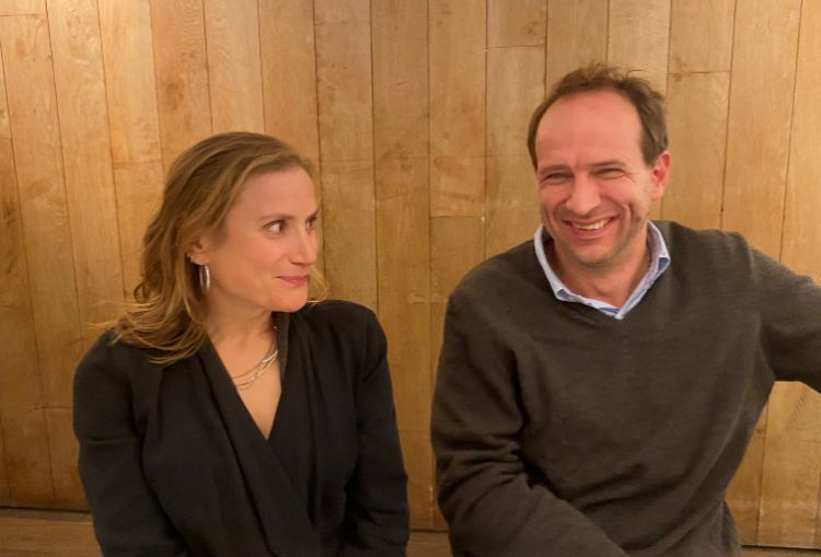 Two people sitting indoors against a wooden panel wall, dressed in smart casual clothing.