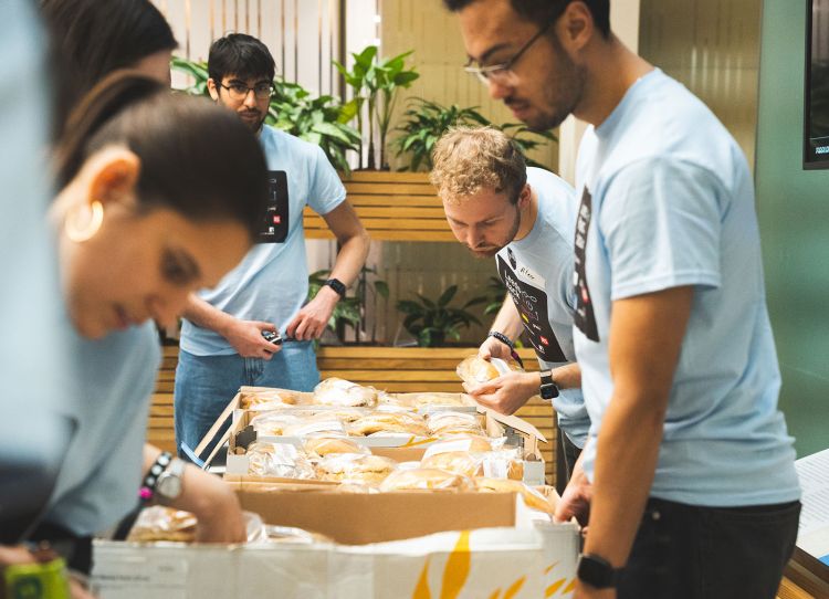 A table filled with sandwiches, surrounded by four people.