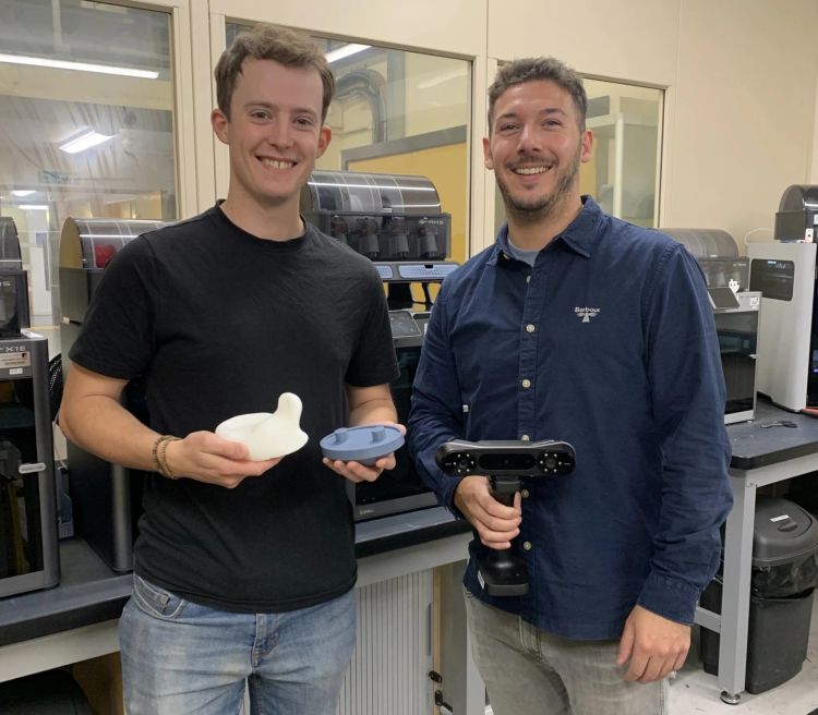 James Watkins and Rhys Moore, standing side by side in a workshop, holding items they have created for REMAP.