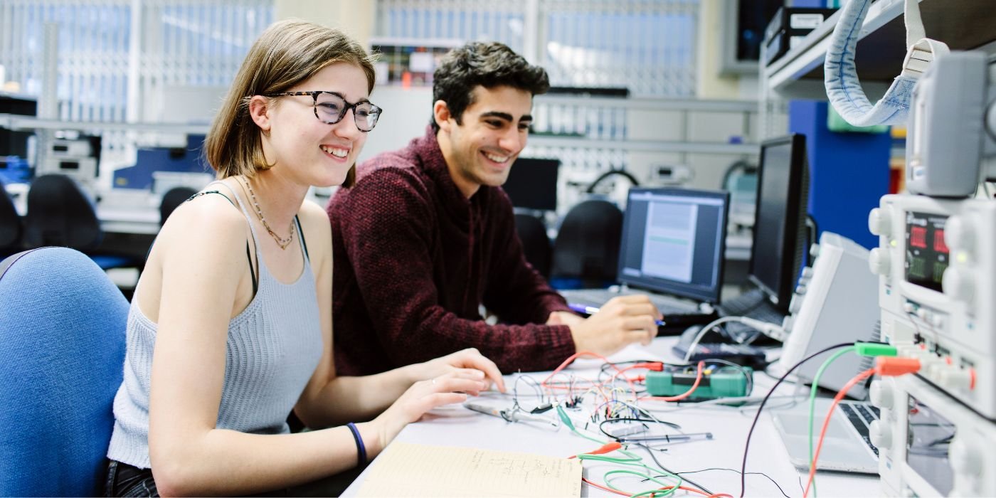 Two students at a work bench in the basement lab in the School of Electronic and Electrical Engineering at the University of Leeds. The students are looking at a piece of equipment with different coloured wires covering the desk.