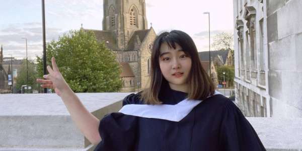 Qianru Yang wearing her graduation gown stands in front of a university building.