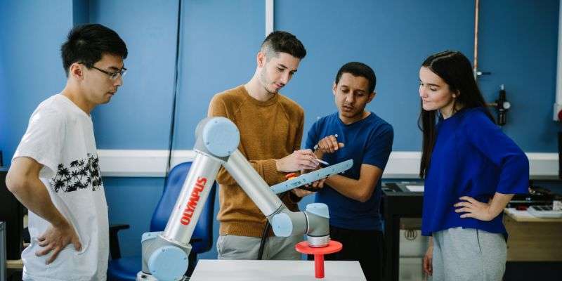 A group of four students are stood around a robotic arm, recording data on a tablet.